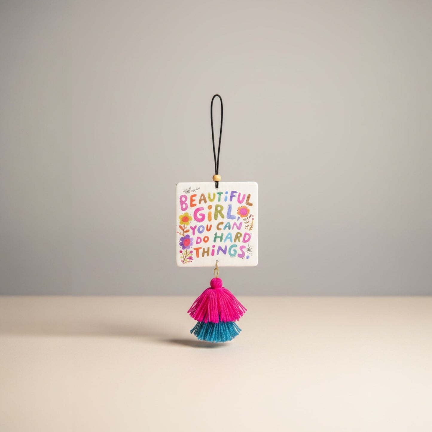 Patchwork with Busyfingers air freshener with colorful text 'BEAUTIFUL GIRL YOU CAN DO HARD THINGS' and floral accents, featuring pink and teal tassels.
