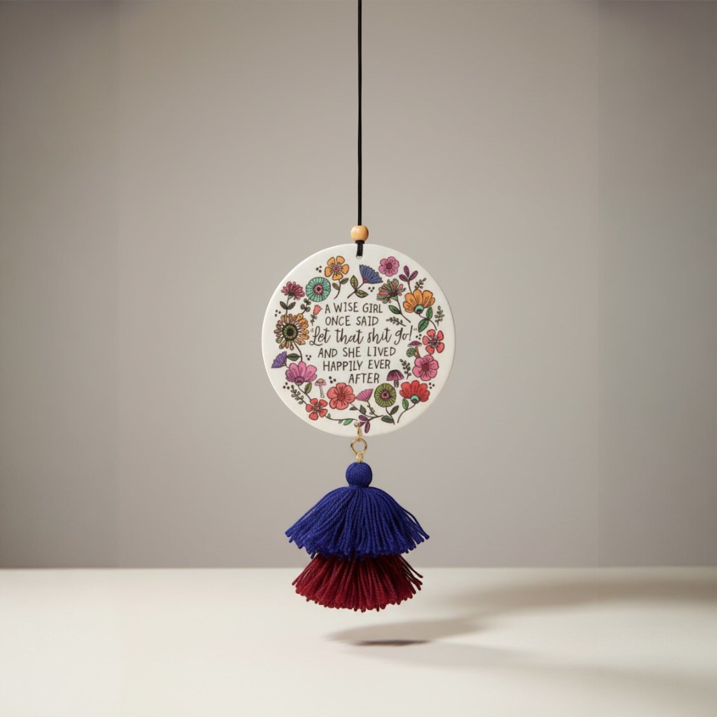 Patchwork with Busyfingers ceramic air freshener with floral design and quote "A wise girl once said let that shit go!" hanging with blue and red tassels.
