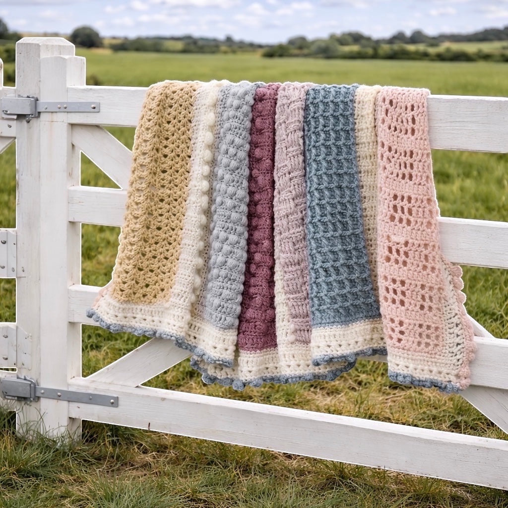 Patchwork with Busyfingers crochet blanket in pastel shades of pink, blue, cream, and mauve, featuring various stitch patterns and bobble details, draped over a white wooden gate.