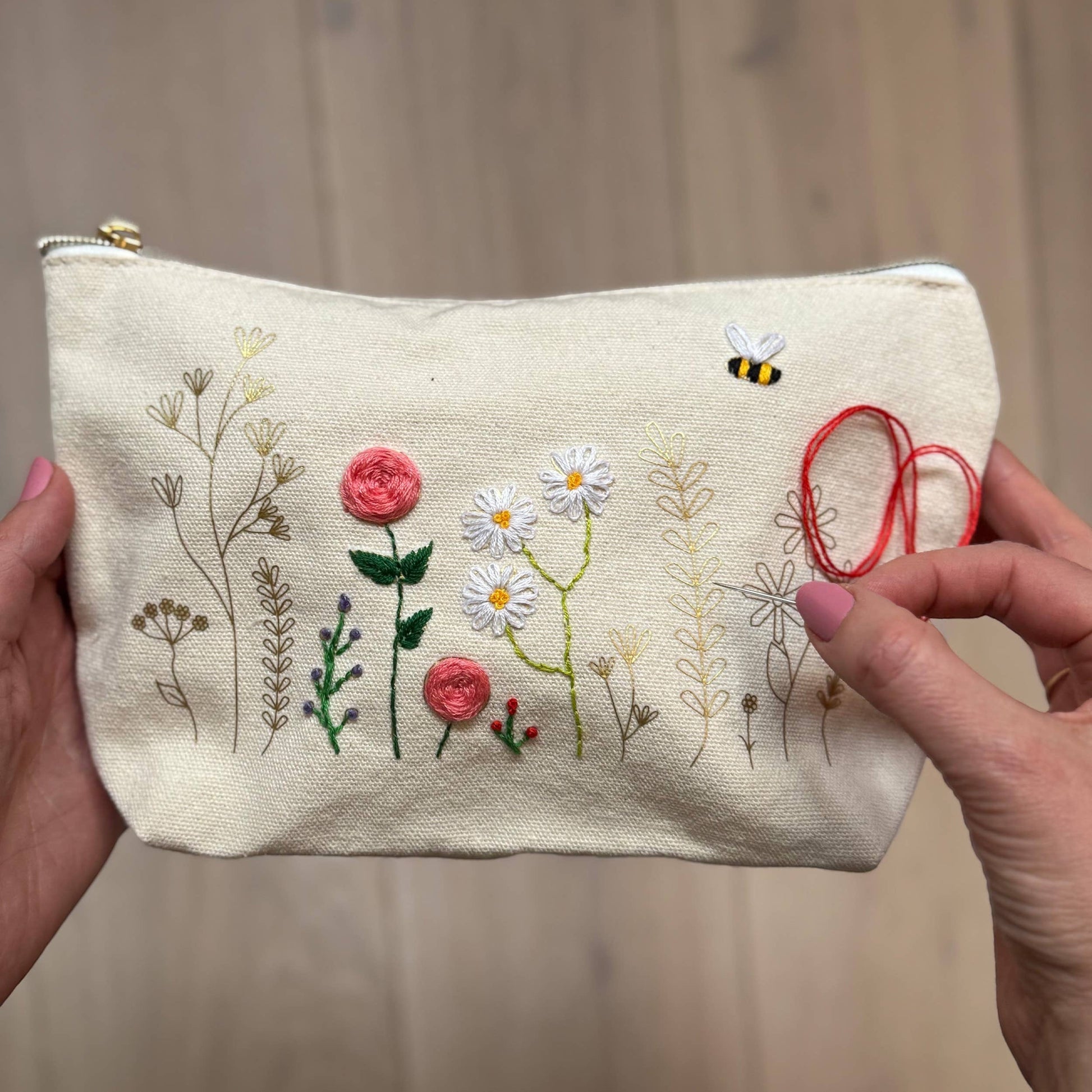Patchwork with Busyfingers floral embroidery kit on a canvas zip pouch with pink roses, daisies, a bee, and red thread detail.