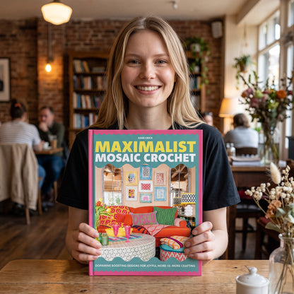 A smiling woman holding the Patchwork with Busyfingers Maximalist Mosaic Crochet book in a cozy cafe setting.